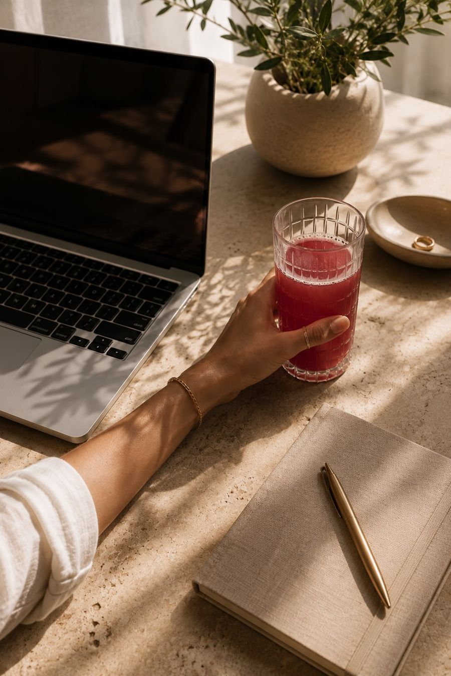 A hand holds a glass of red wellness drink at a sunlit desk beside a laptop and journal